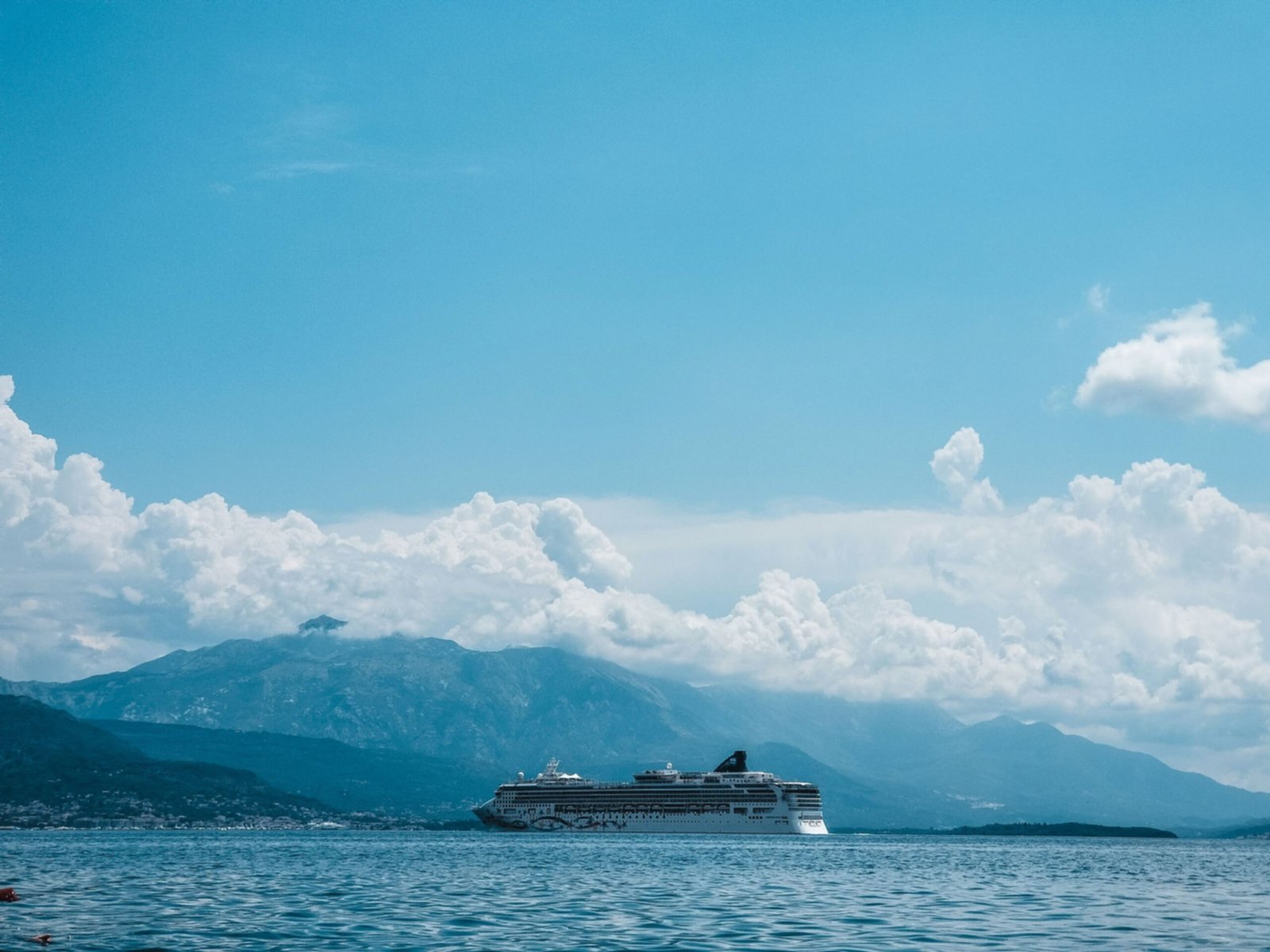 A cruise ship sails across a calm ocean with a backdrop of majestic mountains and clear blue skies.